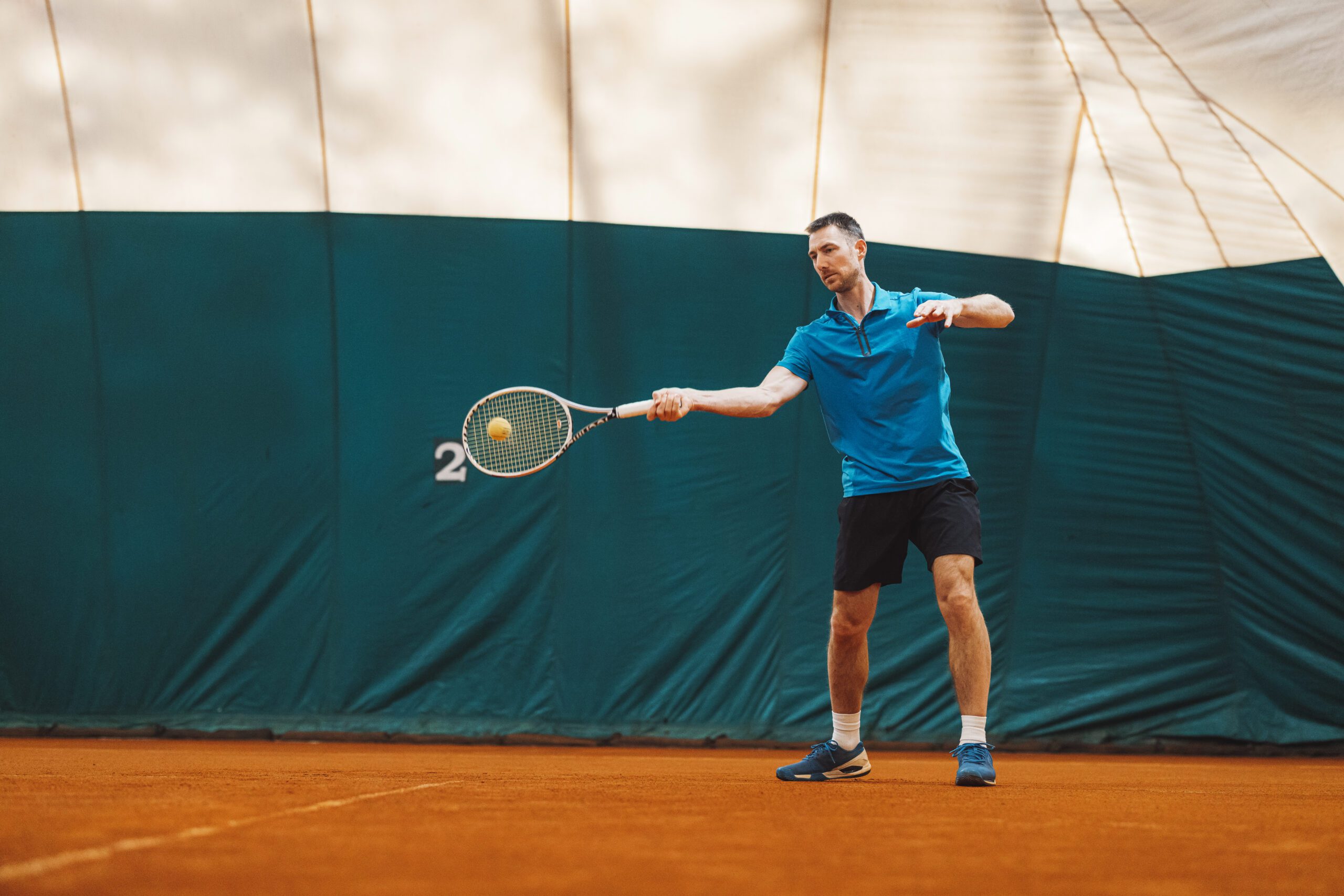 A male tennis player in a blue shirt and black shorts is preparing to hit a backhand shot on an indo